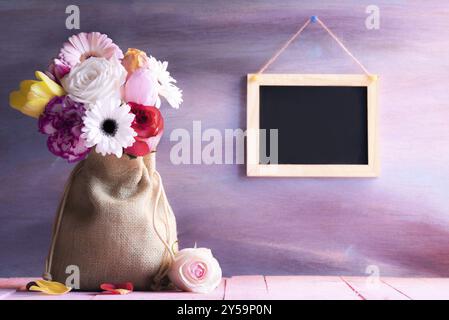 Damentagskarte mit einem Strauß aus Tulpen, Rosen und Gerbera in einer Vintage-Leinwandtasche, auf einem pinken Tisch und einer leeren Tafel auf einer lila Holzwand Stockfoto