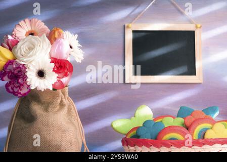 Wunderschöner Blumenstrauß in einem Jutesack, ein Korb mit bunten Keksen und eine leere Tafel, die an der lila Wand hängt Stockfoto