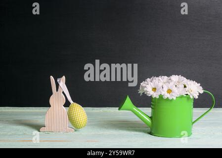 Niedliche weiße Gänseblümchen-Anordnung in einer Gießkanne und ein hölzerner Hase mit einem gelben Stoffei, auf einem grünen Tisch und einem schwarzen Hintergrund Stockfoto