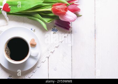 Wunderschöner Blumenstrauß aus bunten Tulpen, gebunden mit Spitze und einer Tasse Kaffee und herzförmigem Zucker, auf einem Vintage Küchentuch, auf einem weißen Holztisch Stockfoto