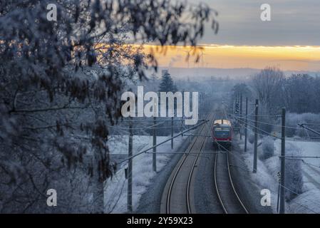 Personenzug fährt auf Bahngleisen durch gefrorene Bäume, bei Sonnenaufgang, in der deutschen Landschaft. Winterreisen. Öffentliche Verkehrsmittel Stockfoto