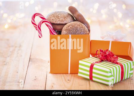 Jede Menge bunte Geschenke, eines voll mit Lebkuchen und Weihnachtsbonbons, auf einem hölzernen Hintergrund, umgeben von verschwommenen Weihnachtslichtern Stockfoto