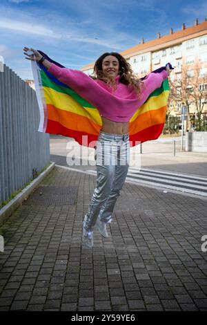 Junge Frau, die mit einer Regenbogenfahne in einer städtischen Umgebung springt Stockfoto