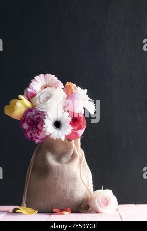 Geschenktüte aus Canvas, gefüllt mit bunten Rosen, Tulpen und Gerbera, auf einem rosafarbenen Tisch und einem schwarzen Hintergrund, umgeben von einer Rose und Blütenblättern Stockfoto