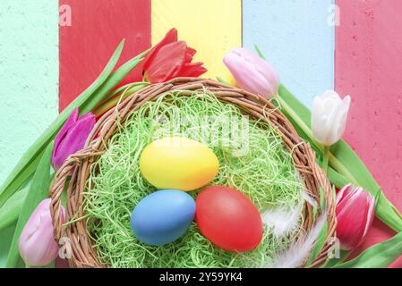 Rustikaler Korb gefüllt mit grünem dekorativem Gras und bemalten Ostereiern, umgeben von wunderschönen Tulpen, auf einem bunten Holzhintergrund Stockfoto