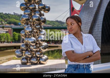 Junge Frau posiert mit überkreuzten Armen in der Nähe der Welpenskulptur und der Zubizuri-Brücke in bilbao, spanien Stockfoto