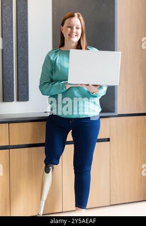 Laptop in der Hand, Frau mit Beinprothese lächelnd im modernen Büro Stockfoto