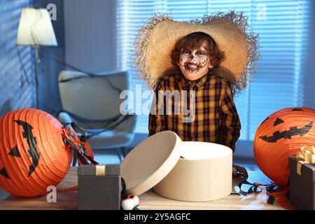 Süßer Junge, der wie Vogelscheuche gekleidet ist, mit festlichem Dekor und Geschenkboxen drinnen in der Nacht. Halloween-Feier Stockfoto