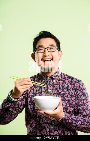 Studio, Porträt und asiatischer Mann mit Essstäbchen für Essen, Ernährung und Lachen mit Selbstvertrauen. Mockup-Raum, männliche Person und Schüssel von Grün Stockfoto