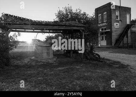 Wenn die Sonne hinter einem verlassenen Brunnen untergeht, wird ein Sonnenstrahl-Effekt sichtbar. Das historische Postamt und die Schotterstraße in einer Geisterstadt. Stockfoto