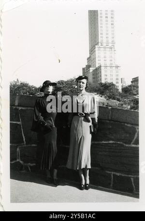 Zwei elegant gekleidete Frauen posieren vor einer Skyline der Stadt und zeigen Mode aus den 1930er Jahren mit strukturierten Kleidern, Gürtelbündchen und stilvollen Hüten. Das Foto fängt die Raffinesse und urbane Eleganz der Epoche ein Stockfoto