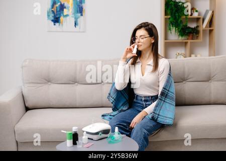 Junge Frau in lässiger Kleidung, mit Nasenspray, um das Atmen zu erleichtern, auf einem Sofa in einem Wohnraum. Stockfoto