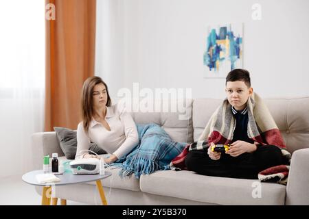 Kranke Dame, die durch Inhalator-Maske zu Hause einatmet, und lustiger Junge mit Gamepad, der Videospiel zu Hause in ihrer Nähe spielt. Stockfoto