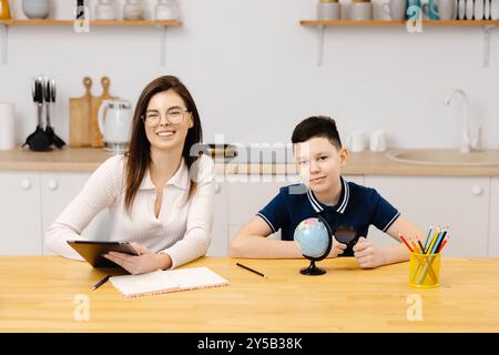 Eine junge Frau hilft einem Jungen beim Unterricht mit einem Tablet und einem Globus. Ein Schüler macht seine Hausaufgaben mit Hilfe eines Tutors. Stockfoto