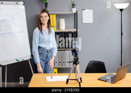 Positive Bloggerin, die auf einem Stativ an der Smartphone-Webcam spricht, einen Beitrag für den Vlog aufnimmt. Online-Lehrer, Coach, Webinar zu Hause. Pressen Stockfoto