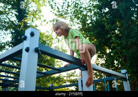 Ein Kind klettert an einem sonnigen Nachmittag souverän auf Spielgeräten unter klarem Himmel, umgeben von Grün Stockfoto