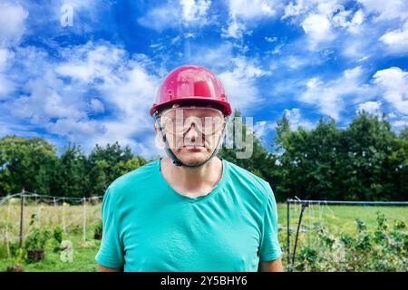 Nahaufnahme eines Baumeisters mit Schutzhelm und Schutzbrille, ein Mann im mittleren Alter, der mit ernstem Ausdruck in die Kamera blickt, vor ländlicher Landschaft, breit Stockfoto