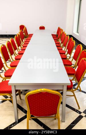 Leerer langer Tisch aus mehreren grauen Holztischen und roten weichen Stühlen in der Halle Stockfoto