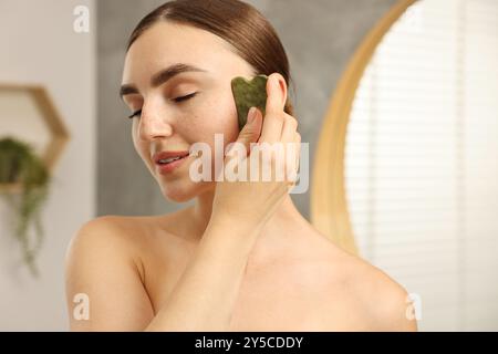 Schöne junge Frau, die eine Gesichtsmassage mit Gua-Sha-Werkzeug drinnen macht Stockfoto