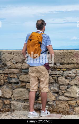 Mann mittleren Alters, der allein auf das Meer blickt, Mann mittleren Alters mit Rucksack allein im Urlaub. Ein Mann, der über eine Mauer am Meer schaut. Stockfoto