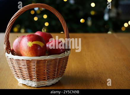 Ein Korbkorb voller roter Äpfel, einer davon mit einem Weihnachtskometen, ein geschmückter, leuchtender Weihnachtsbaum im Hintergrund. Stockfoto