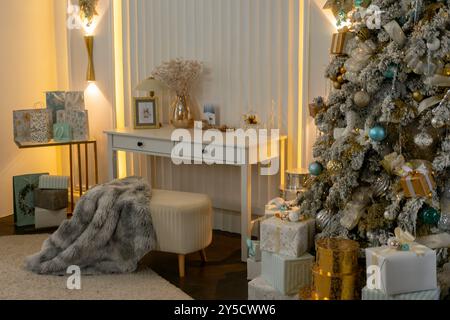 Ein weißer Weihnachtsbaum mit goldenen und blauen Dekorationen steht vor einem weißen Schreibtisch. Der Schreibtisch ist überfüllt mit Geschenken und Geschenken und einer grauen Decke Stockfoto