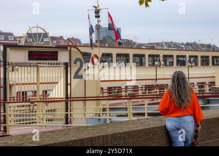 Ältere Frau, die mit dem Rücken zur Kamera auf dem Damm am Eingang Nummer 2 steht, an Bord eines Touristenbootes, Tour auf dem Fluss Maas, niederländische Flaggen, bewölkter Tag i Stockfoto