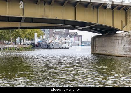 Stahlkonstruktionen am Fuß der Brücke, Schiffe, die am Damm entlang der Maas ankern und Fabriken in unscharfem Hintergrund, bewölkter Tag in Maastricht Stockfoto