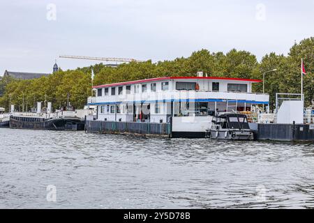 Gruppe von Booten, die am Uferkai entlang des Flusses Maas ankern, grünes Laub von Bäumen im Hintergrund vor grauem Himmel, ruhiges Wasser, bewölkter Tag in Maastrich Stockfoto