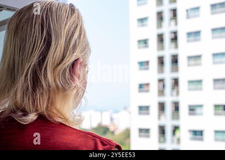 Ein reifer Mann steht auf dem Balkon eines Hauses gegenüber einem hohen Gebäude mit Fenstern. Stockfoto