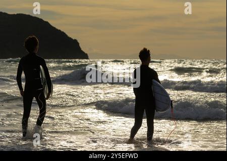 Zwei junge Männer mit Surfbrettern bekämpfen die Wellen bei Sonnenuntergang im Golf von Baratti, Piombino, Livorno, Italien Stockfoto