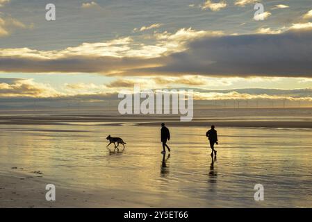 Zwei Personen, die mit einem Hund an einem ruhigen Strand bei Sonnenuntergang spazieren gehen, mit Windrädern in der Ferne. Stockfoto