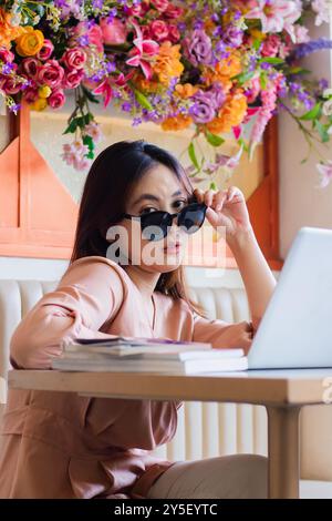 Charme der schönen jungen indonesien Frau elegant zu Kamera hält und trägt Sonnenbrille entspannt auf Sofa im Indoor Coffee Shop sitzt. Für Mode, Stockfoto