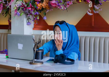 Die Geschäftigkeit eines jungen indonesien-Mädchens, das ein Kopftuch trägt, ist abwesend, wenn er vor einem Laptop, der auf einem Sofa in einer Innenverkleidung sitzt, Büroarbeit verrichtet Stockfoto