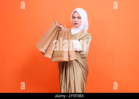 Wütende reife asiatische Frau mit beiden Händen, die eine braune Papiertüte mit Lebensmitteln vor sich trägt, die ein muslimisches Kaftan-Kleid mit einem Hijab trägt Stockfoto