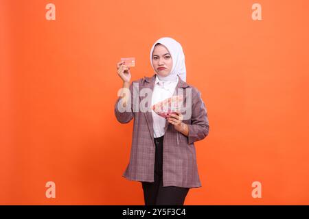 Die junge indonesienfrau in ihren 20ern runzelnd trägt Hijab Bürokleidung, die Debitkreditkarten und Geld hält, isoliert auf einem einfachen, pastellfarbenen Hintergrund stu Stockfoto