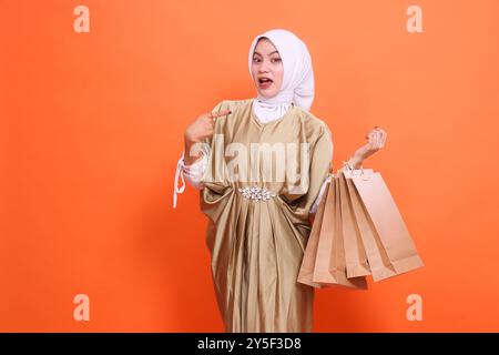 Schockierte reife asiatische Frau, die eine braune Papiertüte mit Einkaufsartikeln trug, während sie darauf zeigte, in einem muslimischen Kaftan-Kleid mit Hijab, auf einem Stockfoto