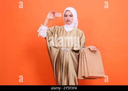 Mürrische junge indonesienfrau mit brauner Papiertüte und Mitgliedskarte hochhaltend, Debitkarte mit Kaftan-Muslimah mit Hijab, isoliert auf OP Stockfoto