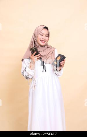 Schönes junges asiatisches muslimisches Mädchen im Hijab, das fröhlich den koran trägt und Gebetskegeln auf dem Smartphone hält. Für Werbung, Lifestyle, Banner und R Stockfoto