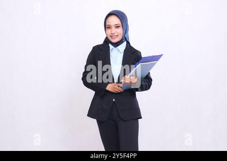 Eine exklusive asiatische Bürofrau mit Hijab steht fröhlich mit Stift, Klemmbrett und Notebook. Schöne muslimische Frau in blauem Hemd Stockfoto