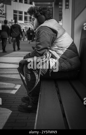 Junger nachdenklicher Mann, der auf einer Bank in einem Stadtpark sitzt und die Musik in seinen Kopfhörern genießt. Zielgerichteter junger Mann mit Kopfhörern in einer Stadt Stockfoto