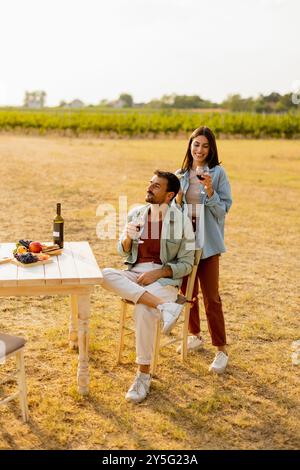 Ein paar entspannen sich an einem rustikalen Tisch in einem sonnigen Weinberg, genießen Wein und Gourmet-Snacks. Sie teilen Lachen und Freude, umgeben von üppigen Weinstöcken und dem Stockfoto