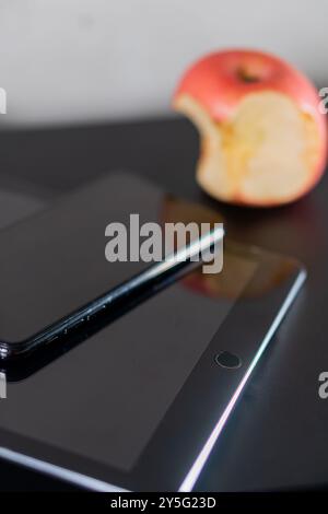 Eine Nahaufnahme eines Smartphones und eines Tablets auf einer schwarzen Oberfläche, mit einem teilweise gegessen roten Apfel im Hintergrund. Die Geräte sind glänzend Stockfoto