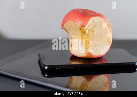 Ein gebissener roter Apfel auf einem schwarzen Smartphone mit reflektierender Oberfläche. Der Hintergrund ist neutral, was den Kontrast zwischen der Frucht und unterstreicht Stockfoto