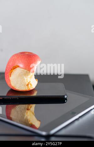 Ein gebissener roter Apfel auf einem schwarzen Smartphone und einem Tablet mit reflektierender Oberfläche. Der Hintergrund ist neutral und minimalistisch, betont Stockfoto