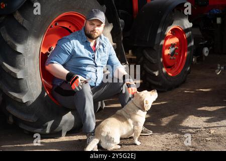 Ein Landwirt repariert Landmaschinen. Er sitzt neben dem Traktor, mit einem gelben Hund neben ihm. Stockfoto