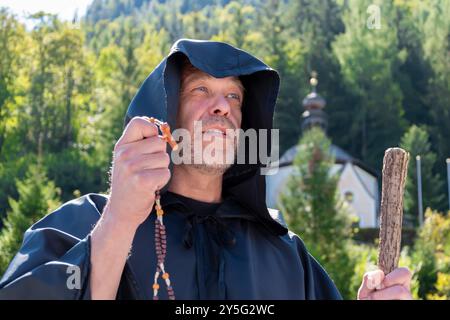 к ein älterer Mönch mit Bart und Personal in dunklen Gewändern hält einen Gebetsrosar, während er vor einem verschwommenen Hintergrund von Bergen und einer Kirche steht. Stockfoto