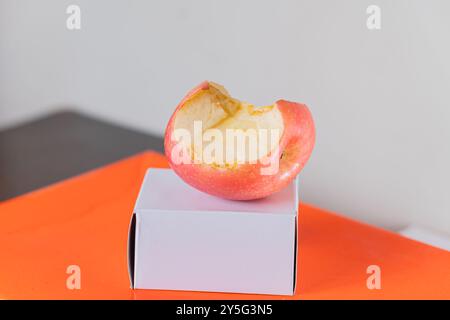 Ein teilweise gegessen roter Apfel, der auf einer kleinen weißen Box liegt und auf einer leuchtend orangefarbenen Oberfläche liegt. Der Apfel zeigt eine Bissspur, die sein hellgelbes Licht offenbart Stockfoto