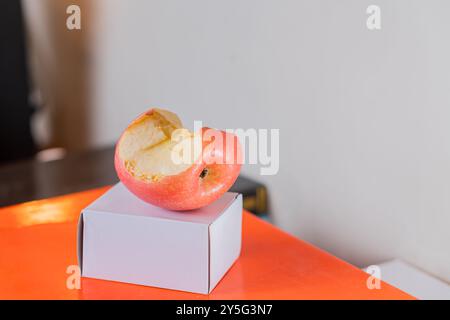 Ein gebissener roter Apfel auf einer kleinen weißen Box, auf einer orangefarbenen Oberfläche. Der Hintergrund ist unscharf und betont den Apfel und die Box. Stockfoto