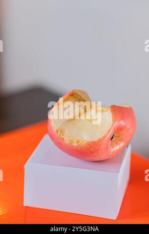 Ein gebissener Apfel, der auf einer kleinen weißen Box mit einer orangefarbenen Oberfläche liegt. Der Apfel ist rosa-rot und hat einen Biss herausgenommen, der sein helles Inneres offenbart. Das b Stockfoto